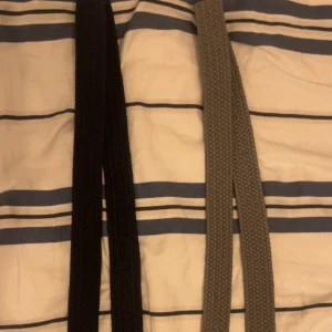 Två flätade herrbälten med läderdetaljer - Säljer två stilrena flätade bälten, ett svart och ett grått. Båda har bruna läderdetaljer vid spännet och änden samt metallspänne. Perfekta för att lyfta din outfit med en klassisk touch.