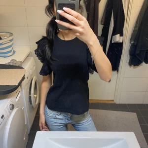 Svart blus med volangärmar - Säljer en svart blus med korta volangärmar i broderad spets. Toppen har en rund halsringning och är i en rak, normal passform. Perfekt för dig som gillar en söt men stilren look.