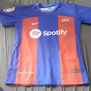 FC Barcelona Lewandowski matchtröja - Säljer en officiell FC Barcelona matchtröja från Nike med Lewandowski och nummer 9 på ryggen. Tröjan har klassiska blå och röda ränder, klubbmärke på bröstet, Spotify-logga och Dri-FIT ADV-material som andas. Kortärmad modell med tryckta detaljer och La Liga-märke på ärmen.