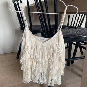Beige fransad topp  - Supersnygg beige topp från NA-KD med tunna axelband och flera lager fransar som ger en riktigt cool och festlig look. 