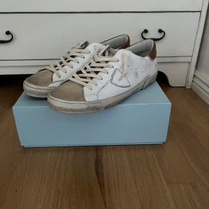 Philippe Model vita sneakers i skinn - Snygga vita sneakers från Philippe Model med beige detaljer och brunt parti vid hälen. Skorna har klassisk snörning, rund tå och är tillverkade i skinn med slitna detaljer för en cool, urban look. Perfekta sommar skorna. Använda 2 gånger, äkta. köpta på Mertch of Sweden.