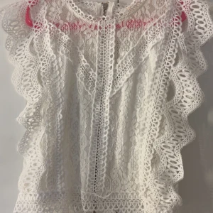 Vit spetsblus med volangdetaljer - Superfin vit blus i spets med vackra volangdetaljer längs axlarna och ärmlös design. Blusen har rund halsringning och dragkedja bak. Perfekt för dig som gillar romantisk och detaljerad stil.