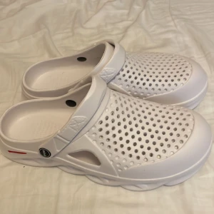 Vita sportiga sandaler med hålmönster - Vita sandaler i syntetmaterial med sportig look och ventilerande hålmönster över hela ovandelen. Justerbar hälrem och rejäl, mönstrad sula för extra grepp. Perfekta för chill dagar eller när du vill ha något bekvämt och luftigt på fötterna.