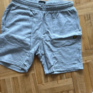 Grå mjuka shorts från Lyle & Scott - Grå shorts från Lyle & Scott med elastisk midja och sidofickor. Mjukt bomullsmaterial och klassisk passform, perfekt för chill dagar. Liten gul logga på ena benet och en bakficka för extra stilpoäng.