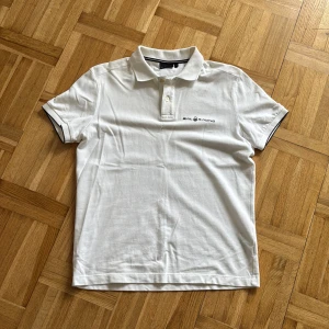 Vit pikétröja från Sail Racing S - Klassisk vit pikétröja från Sail Racing i storlek S. Tröjan har korta ärmar, krage med knappar och svart logga på bröstet. Snygga svarta detaljer på krage och ärmslut. Perfekt till sommaren och enkel att matcha med jeans eller shorts.