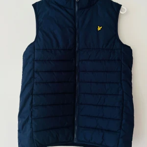 Mörkblå dunväst från Lyle & Scott Junior - Snygg mörkblå dunväst från Lyle & Scott Junior med gul logga på bröstet. Västen har quiltad design och dragkedja framtill. 