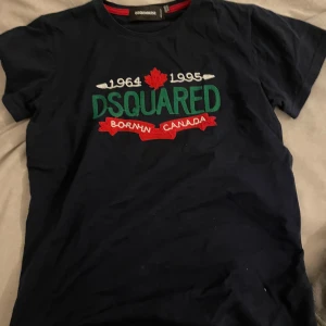 Mörkblå t-shirt från Dsquared2 - Snygg mörkblå t-shirt från Dsquared2 med broderad text och lönnlöv på bröstet. Texten 'DSQUARED', '1964 1995' och 'Born in Canada' i grönt, vitt och rött. Perfekt för dig som gillar streetwear och vill sticka ut.