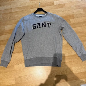 Grå sweatshirt från GANT - Klassisk grå sweatshirt från GANT med rund halsringning och stora broderade bokstäver på bröstet. Tröjan har ribbade muddar vid ärmslut och nederkant. Perfekt för en avslappnad och stilren look. Skicket är väldigt bra. Storlek M
