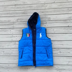 Ralph Lauren väst - Snygg blå dunväst med huva från Polo Ralph Lauren. Västen har broderad logga på bröstet, italiensk flagga och texten 'ITALIA' samt nummer 6 på ryggen. Två fickor med dragkedja framtill och quiltad design.