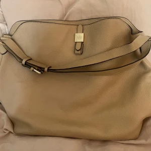Beige axelväska från Mulberry - Säljer en klassisk beige axelväska från Mulberry i skinn med guldfärgade metalldetaljer och präglad Mulberry-logga. Väskan har en bred axelrem och  ett elegant spänne framtill. Perfekt för dig som gillar stilrena accessoarer.