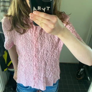 Rosa kortärmad skjorta/blus från Lindex - Säljer en söt rosa skjorta/blus från Lindex i storlek S. Den har korta ärmar, krage och knappar framtill. Materialet är krinklat och ger en cool textur. Perfekt till jeans eller kjol för en fräsch look! Nästan aldrig använd så i väldigt bra skick!