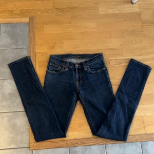 Mörkblå jeans från Nudie Jeans - Säljer ett par mörkblå jeans från Nudie Jeans med klassisk femficksdesign och orangea sömmar. Byxorna har normal passform och raka ben. Nudie-loggan syns på bakfickan och läderpatchen i midjan.
