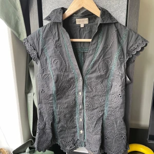 Svart broderad blus från Karen Millen - Säljer en svart blus från Karen Millen med broderade blommor och dekorativa spetsdetaljer. Blusen har korta ärmar med vågig kant, knappar framtill och en feminin krage. Perfekt för dig som gillar unika detaljer och snygg passform.