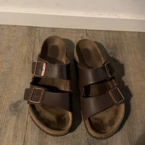 Bruna sandaler från Birkenstock - Klassiska bruna sandaler från Birkenstock med två breda remmar i skinn och justerbara metallspännen. Fotbädden är anatomiskt utformad för extra komfort och sulan är i kork. Perfekta för sommardagar och enkel att matcha med det mesta. OBS SKORNA KOMMER SANDALERNA KOMMER SÅKLART FINA OCH TVÄTTADE!