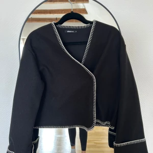 Svart omlottjacka från Gina Tricot - Svart omlottjacka från Gina Tricot med kontrasterande vita sömmar längs kanterna. Jackan är croppad och har lång ärm, perfekt för att lyfta en enkel outfit. Stilren och trendig design med markerade detaljer.