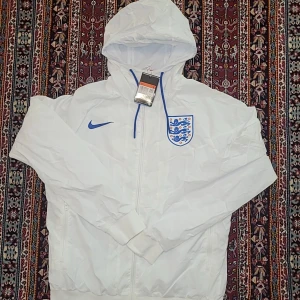 Vit England Nike vindjacka med huva - Snygg vit vindjacka från Nike med Englands landslagsmärke på bröstet och blå Nike-logga. Jackan har huva, dragkedja framtill och meshfoder för extra komfort. Perfekt för fotbollsfans som vill ha en clean och sportig stil.