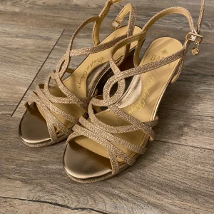 Glittriga guldiga sandaler från Dasia - Snygga klackskor från Dasia i guld med glittriga remmar och öppen tå. Skorna har justerbart spänne vid ankeln. Perfekta för dig som vill sticka ut med en elegant touch. Strappy design och metallic finish ger en lyxig känsla.