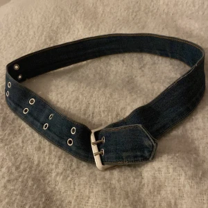 Snyggt jeansbälte med dubbla spännen - Snyggt och brett jeansbälte i mörkblå denim med dubbla metallspännen och silverfärgade öljetter. Perfekt accessoar för att ge din outfit en cool och avslappnad vibe. Bältet har markerade sömmar och en spetsig avslutning framtill. Perfekta till ett par snygga jeans!