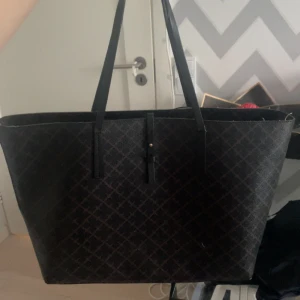 Svart väska By Malene Birger - Stilren svart shoppingväska från By Malene Birger med klassiskt rutigt mönster och långa axelband. Väskan är rymlig med beige insida och har en praktisk rem med spänne för nycklar eller småsaker. Perfekt för dig som vill ha plats för allt du behöver under dagen. 