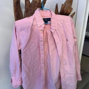 Rosa skjorta från Ralph Lauren - Snygg rosa skjorta från Ralph Lauren med klassisk krage och knappar framtill. Skjortan har lång ärm och ikonisk broderad logga på bröstet. Tillverkad i mjuk bomull som känns skön mot huden. Perfekt för dig som gillar stilrena och tidlösa plagg. behövs bara stryckas lite. Storlek xs