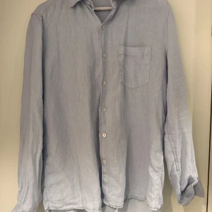 Ljusblå linneskjorta från A Day’s March - Snygg ljusblå skjorta i linne med klassisk krage och knäppning framtill. Skjortan har en bröstficka och långa ärmar med manschett. Perfekt för en avslappnad men stilren look.