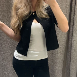 Svart kort kofta med knapp - Snygg svart kort kofta med en dekorativ guldig knapp upptill. Koftan har korta ärmar och en öppen front, perfekt att slänga över ett linne eller t-shirt. Enkel och stilren design som passar till många outfits.