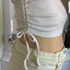 Vit oneshoulder croppad topp  - Säljer en vit ribbad oneshoulder topp från newyorker. Toppen är croppad och har snörning i sidan med en rosett. Perfekt för varma dagar i sommar.