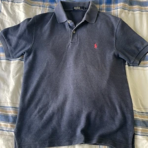 Mörkblå pikétröja från Polo Ralph Lauren - Säljer en klassisk mörkblå pikétröja från Polo Ralph Lauren med röd broderad logga på bröstet. Tröjan har krage, knappar vid halsen och är tillverkad i 100% bomull. Perfekt för en stilren sommar look eller varför inte ute på ranchen. Priset går att diskuter!