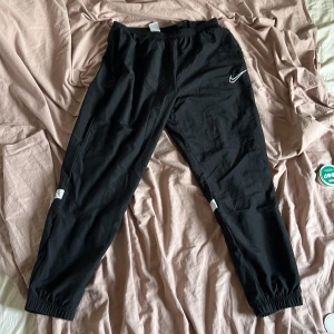 Svarta träningsbyxor från Nike med vita detaljer - Svarta Nike Dri-Fit träningsbyxor med vita reflexdetaljer på benen och resår i midja och benslut. Perfekta för sport eller chill, med klassisk Nike-logga på låret och bekväm passform.