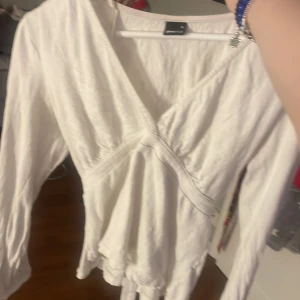 Vit omlottblus med knyt - Säljer en vit omlottblus med knyt i midjan och v-ringning. Blusen har långa ärmar och är tillverkad i ett lätt, lite glansigt material.  Säljer då den ej används längre! 