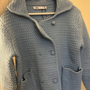 Ljusblå stickad jacka från Zara - Mysig ljusblå stickad jacka från Zara med stora knappar framtill och två fickor. Jackan har lång ärm och en bred krage, perfekt för lager på lager under kyliga dagar.