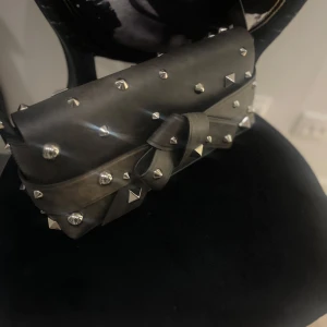 Svart handväska med nitar från Acne Studios - Cool svart handväska från Acne Studios med silverfärgade nitar och en stor rosett framtill. Väskan har ett handtag och är tillverkad i skinn. Perfekt för dig som vill sticka ut med en edgy accessoar.