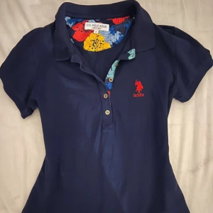 Mörkblå pikétröja från U.S. Polo Assn. - Säljer en mörkblå pikétröja från U.S. Polo Assn. med klassisk krage och knappar framtill. Insidan av kragen har ett färgglatt blommigt mönster och loggan är broderad i rött på bröstet. Perfekt för en sportig och stilren look.