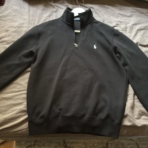 Svart halvzip sweatshirt från Polo Ralph Lauren - Klassisk svart sweatshirt från Polo Ralph Lauren med halv dragkedja och broderad logga på bröstet. Tröjan har lång ärm och ribbade muddar. Perfekt för en stilren och avslappnad look. Som ny kostar den 2200kr