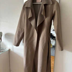 Beige kappa från Nelly - Säljer en stilren beige kappa från Nelly med lång passform och klassisk krage. Jackan har långa ärmar och ett bälte i midjan för att justera. Lite nopprig därav priset 💕