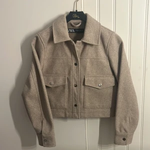 Beige croppad jacka från Zara - Stilren beige jacka från Zara med croppad passform och klassisk krage. Jackan har två bröstfickor med lock och knappar samt knäppning framtill. Perfekt för lager-på-lager och enkel att matcha.
