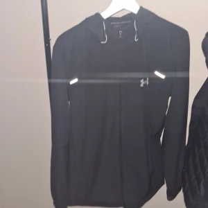 Svart vindjacka från Under Armour - Säljer en svart vindjacka från Under Armour med huva och dragkedja. Jackan har reflekterande detaljer och logga på bröstet samt tryckt logga på ryggen och ärmslutet. Perfekt för träning utomhus under blåsiga dagar.