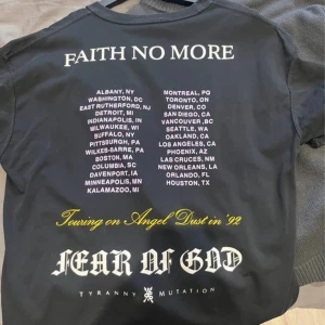 Svart Faith No More t-shirt med turnétryck - Säljer en svart t-shirt från Fear of God med Faith No More-turnétryck på ryggen. T-shirten har vit och gul text samt detaljerade tryck med städer och logotyper. Perfekt för dig som gillar bandmerch och streetwear.
