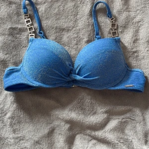Blå glittrig bikiniöverdel med dekorativa band - Säljer en blå bikiniöverdel med glittrigt tyg och snygga silverfärgade detaljer på axelbanden. Överdelen har vadderade kupor och knytdetalj framtill. Perfekt för stranden eller poolen!