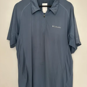 Blå funktionspiké från Columbia - Säljer en blå funktionspiké från Columbia med halv dragkedja och korta ärmar. Perfekt för träning eller utomhusaktiviteter. Materialet är lätt och andas bra. Diskret logga på bröstet och sportig design.