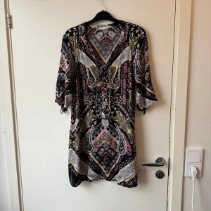 Mönstrad klänning med knyt i midjan - Säljer en snygg mönstrad klänning med v-ringning och knyt i midjan. Klänningen har trekvartsärm och ett färgstarkt mönster i svart, rosa, gul och vit. Perfekt för dig som gillar bohemiska vibes och unika prints. Från märket Odd Molly i storlek 0 som enligt deras storleksguide motsvarar ungefär en Storlek XS. Fint skick, men kan vara någon liten tråddraning. 