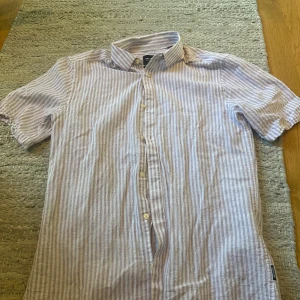 Vit och ljuslila randig skjorta från Only & Sons - Säljer en fräsch skjorta från Only & Sons i vit och ljuslila med smala ränder. Skjortan har korta ärmar, klassisk krage och knäppning framtill. Perfekt för varma dagar och stilrena outfits.