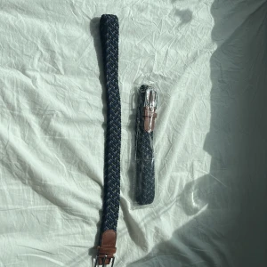 Flätat blått och grått bälte med skinn - Snyggt flätat bälte i blått, svart och grått med detaljer i brunt skinn och silverfärgat spänne. 