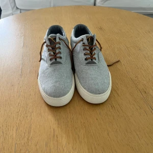 Polo Ralph Lauren skor - Snygga grå sneakers från Polo Ralph Lauren med vit sula och bruna snören. Skorna är i textil med diskret logga på sidan och POLO-broderi bak. Perfekta för en clean och avslappnad stil.