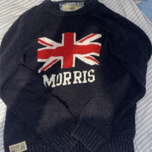 Mörkblå stickad tröja Morris S - Snygg mörkblå stickad tröja från Morris med Union Jack-flagga och logga framtill. Tröjan har rund hals, långa ärmar och ribbade muddar. Perfekt för dig som gillar klassisk brittisk stil och vill sticka ut lite extra.