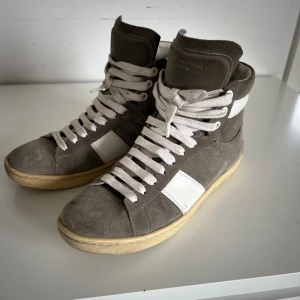 Grå höga sneakers från Saint Laurent - Säljer ett par grå höga sneakers från Saint Laurent Paris med vita detaljer och vit sula. Skorna har snörning framtill och är tillverkade i mocka med inslag av vitt skinn. Klassisk och stilren design med rund tå. Använda men i bra skick❤️