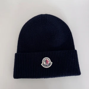Mörkblå ribbad mössa från Moncler - Snygg mörkblå ribbstickad mössa från Moncler med uppvikt kant och broderad Moncler-logga framtill. Perfekt för kyliga dagar och enkel att matcha med olika outfits.