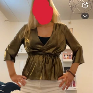 Guldig omlottopp med trekvartsärm - Säljer en glansig guldig omlottopp med trekvartsärm och markerad midja. Toppen har en snygg v-ringning och ett elegant omlottband i midjan. Perfekt för dig som vill sticka ut lite extra!