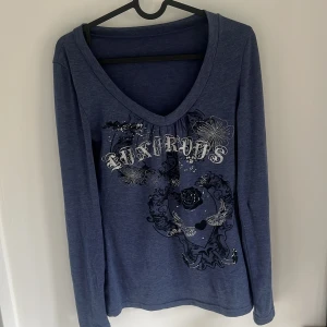 Blå långärmad topp med tryck - Säljer en blå långärmad topp med djup v-ringning och tryck i vitt och svart framtill. Texten 'LUXURIOUS' och blommönster ger en cool och unik look. Perfekt för dig som vill sticka ut lite extra.
