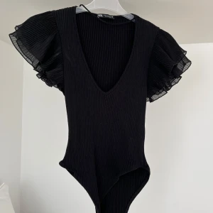 Svart ribbad bodysuit med volangärmar från Zara - Säljer en svart ribbad bodysuit från Zara med djup v-ringning och korta volangärmar i plisserat tyg. Perfekt för att lyfta vilken outfit som helst med sin stilrena och trendiga look.